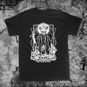Könnte beinhalten: Schwarzes T-Shirt mit einem weißen Grafikdruck eines Katers, der eine Robe trägt und einen Zauberstab hält. Der Kater ist von Schädeln umgeben und dem Text "THE MAGICIAN".
