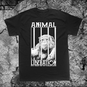 Może przedstawiać: Czarny T-shirt z białym nadrukiem przedstawiającym szympansa w klatce. Tekst "ANIMAL LIBERATION" znajduje się nad obrazem, a "ZOO CAGES ARE NOT CONSERVATION" pod nim.