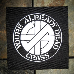 May include: Black and white embroidered patch with the text "You're Already Dead" and a circle with a cross inside. The circle has the text "CRASS" around it.