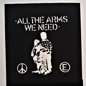May include: Black and white patch with the text "ALL THE ARMS WE NEED." and a white silhouette of a person holding a peace sign. There is a peace sign and a letter "E" on either side of the person.