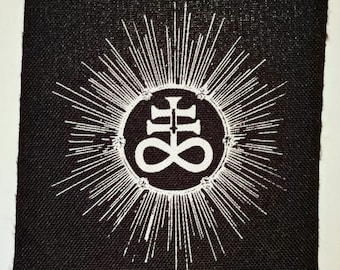 Könnte beinhalten: Ein schwarzer Stoff-Patch mit einem weißen Symbol eines Kreuzes mit einem Unendlichkeitszeichen in einem Kreis, umgeben von einem Sonnenstrahlendesign.