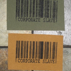 May include: Two fabric patches with a black barcode and the text "CORPORATE SLAVE" printed on them. The patches are on a brown and gray background.