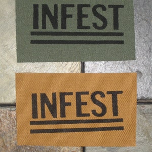Pode incluir: Dois patches de tecido com a palavra "INFEST" impressa em letras pretas. Os patches são feitos de materiais diferentes, um é verde e o outro é castanho.