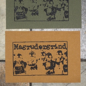 May include: Two brown fabric patches with black printed text and images. The text on both patches reads "Magrudergrind". The image on each patch shows a group of three people with their faces obscured.