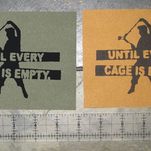 May include: Two fabric patches with the text "UNTIL EVERY CAGE IS EMPTY." printed in black. The patches are on a brown surface. One patch is green and the other is orange.