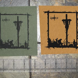 May include: Two square patches with black outlines and black figures inside. The patches are made of a brown fabric and a green fabric. The figures are of a person hanging from a cross with other figures at the base of the cross. The text "Aphex Twin" is printed on the patches.