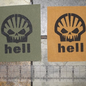 Puede incluir: Dos parches de tela con un diseño de calavera y huesos cruzados negros y la palabra "hell" impresa debajo. Los parches están sobre una superficie marrón con una regla para la escala.