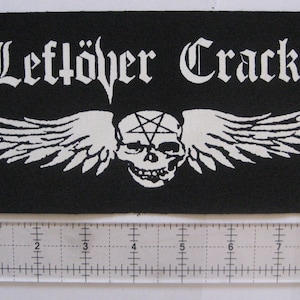 May include: Black patch with the words "Leftöver Crack" in gothic font above a skull with wings and a pentagram. The skull and wings are white. A ruler is placed below the patch.