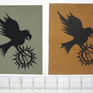 May include: Two fabric patches with a black silhouette of a bird holding a circular symbol with the letters "AF" inside. The patch on the left is on a green background and the patch on the right is on a brown background.