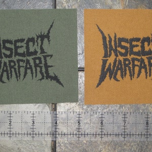 May include: Two black and white embroidered patches with the text "Insect Warfare" on a green and brown fabric background.