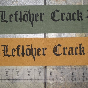 Può includere: Due toppe di tessuto con lettere nere e un pentagramma. La toppa superiore è verde e la toppa inferiore è arancione. Il testo su entrambe le toppe recita "Leftöver Crack".