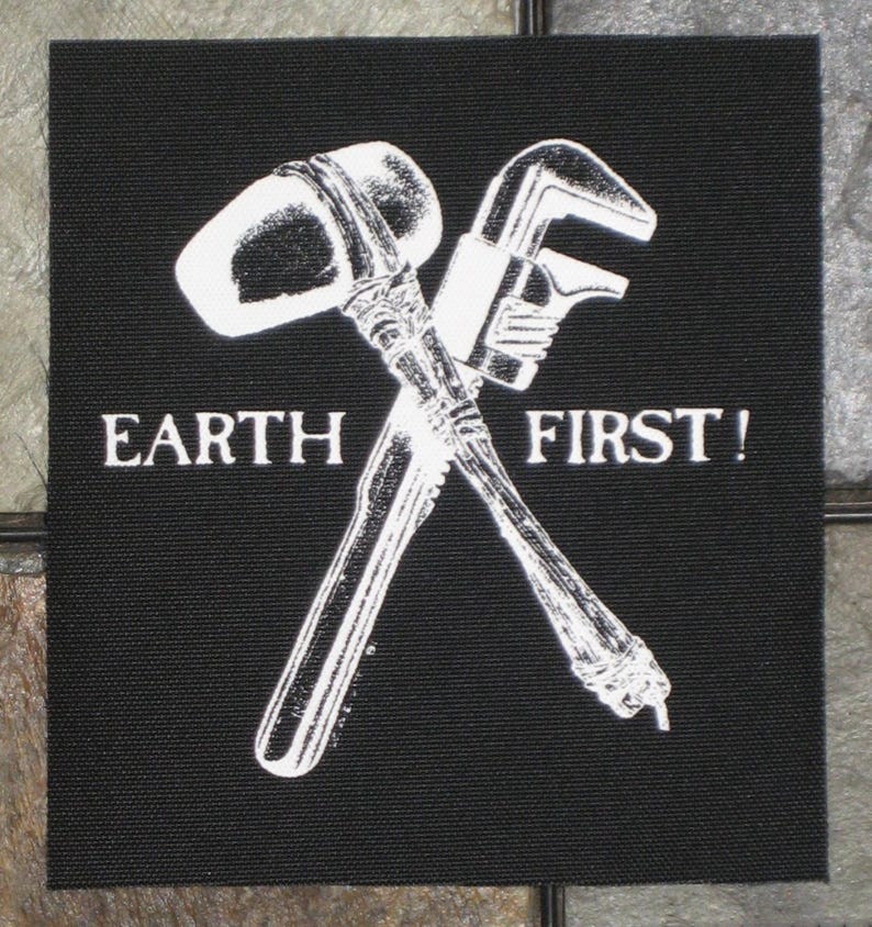 May include: Black and white patch with a crossed hammer and wrench. The text "EARTH FIRST!" is printed below the tools.