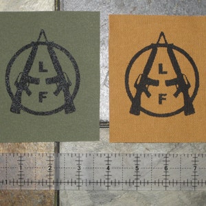 Puede incluir: Dos parches de tela con el mismo diseño. El diseño es un círculo negro con dos rifles estilizados cruzados en su interior. Las letras "L" y "F" están en el centro de los rifles. Un parche es verde oliva y el otro es tostado.