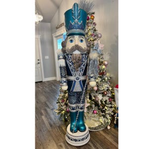 Puede incluir: Una gran estatua decorativa de cascanueces en tonos turquesa, azul marino y plateado. El cascanueces tiene un sombrero alto, un bigote largo y sostiene un bastón. La estatua mide aproximadamente 183 cm de altura y se apoya sobre una base decorativa. Un árbol de Navidad está en el fondo.