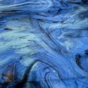 May include: Abstract glass art piece with a flowing design in shades of blue, white, and black. The colors blend and swirl, creating a dynamic, textured appearance. The piece evokes a sense of movement and depth.