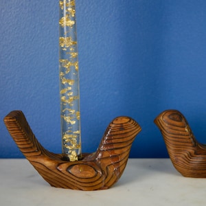 May include: Two wooden bird-shaped candle holders with clear glass tubes filled with gold flakes. The holders are a dark brown color and have a natural wood grain.