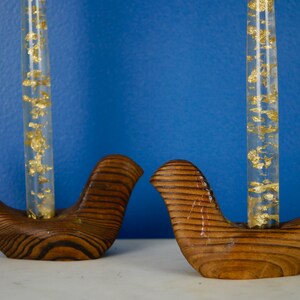 May include: Two wooden bird-shaped candle holders with clear glass tubes filled with gold flakes. The holders are brown with a wood grain pattern.