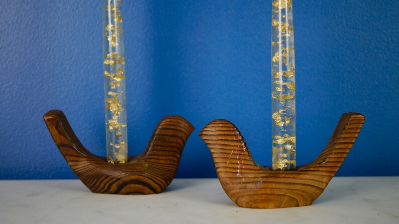 May include: Two wooden bird-shaped candle holders with clear glass tubes filled with gold flakes. The holders are brown with a wood grain pattern.