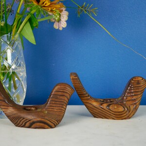 May include: Two wooden bird candle holders with a natural wood grain finish. The holders are in the shape of birds with their wings outstretched.