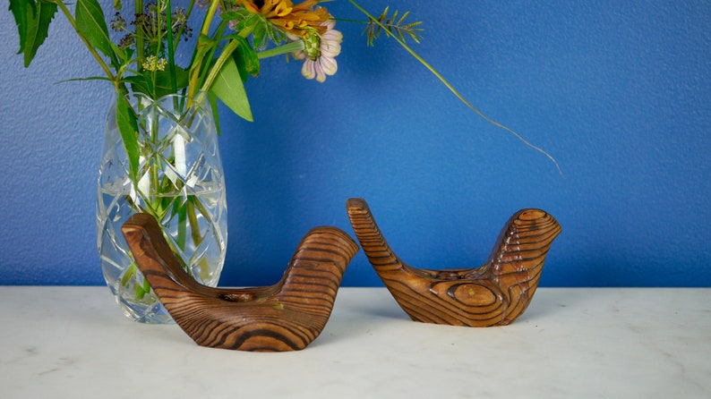 May include: Two wooden bird candle holders with a natural wood grain finish. The holders are in the shape of birds with their wings outstretched.