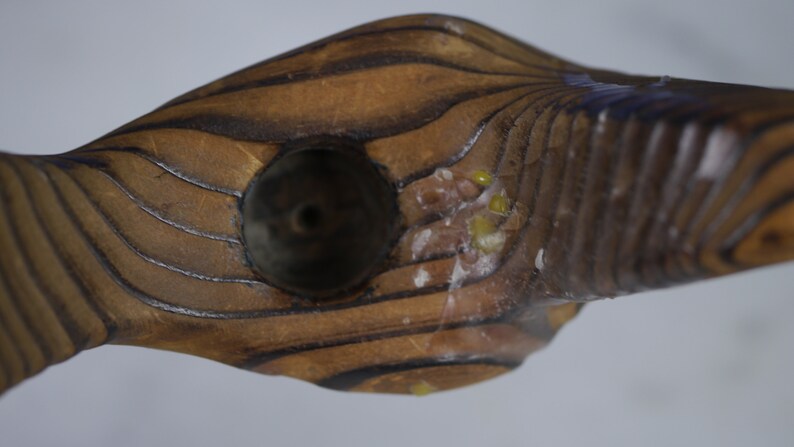 May include: A wooden pipe with a dark brown finish and a round bowl. The bowl has a small hole in the center and is surrounded by a light brown ring. The pipe is decorated with a series of lines that run along the length of the pipe.
