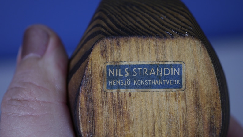 May include: A wooden sculpture with a blue label that reads "NILS STRANDIN HEMSJ&Ouml; KONSTHANTVERK".