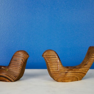 May include: Two wooden bird candle holders in a light brown color. The birds are facing each other and have a slightly curved shape.