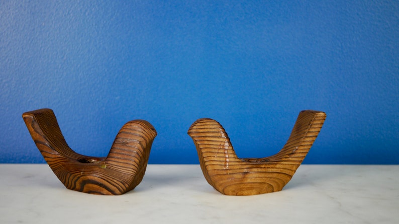 May include: Two wooden bird candle holders in a light brown color. The birds are facing each other and have a slightly curved shape.