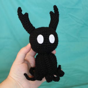 May include: A handmade black crochet creature with white eyes and two curved antennae. The plush toy has a rounded body and small tentacles. The toy is held against a teal background.