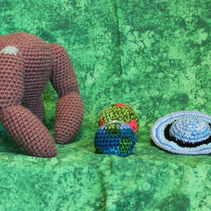 May include: A brown crocheted creature with four thick limbs, two small round crocheted spheres in green, red, and blue, and a flat, circular crocheted hat featuring concentric rings of light blue, black, and white yarn.
