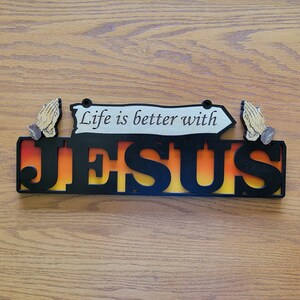May include: A wooden sign with the words "JESUS" in large, black, cut-out letters, with an orange and yellow gradient background. Above the word "JESUS" is the phrase "Life is better with" on a white arrow-shaped sign. Two praying hands are on either side.