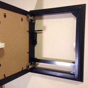 May include: A black framed cabinet door, open to reveal a tan backing. The door is attached to the frame with silver hinges. The cabinet is mounted on a beige wall.