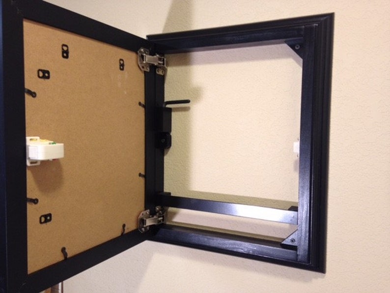 May include: A black framed cabinet door, open to reveal a tan backing. The door is attached to the frame with silver hinges. The cabinet is mounted on a beige wall.