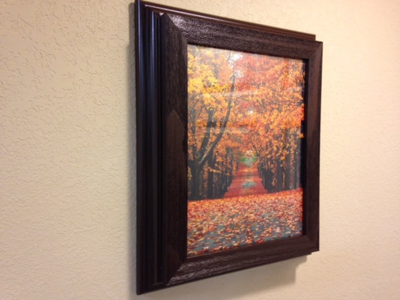 May include: A framed print of a fall scene. The image features a path lined with trees with vibrant orange and yellow leaves. The frame is dark brown with a textured finish, adding a classic touch to the artwork.