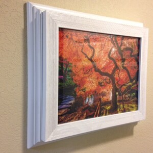 May include: A framed print of a Japanese maple tree with vibrant red and orange leaves. The artwork is encased in a white, textured frame, mounted on a beige wall. The image captures the tree's intricate branches and the sunlight filtering through the foliage.