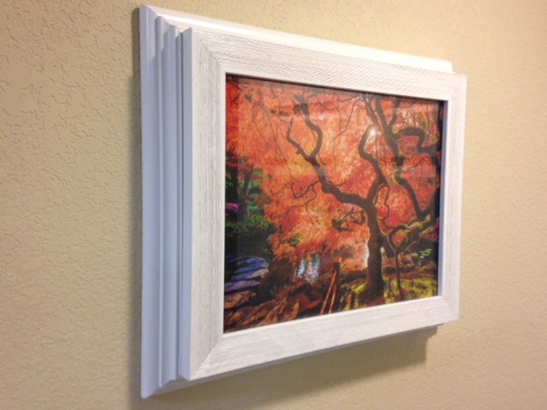 May include: A framed print of a Japanese maple tree with vibrant red and orange leaves. The artwork is encased in a white, textured frame, mounted on a beige wall. The image captures the tree's intricate branches and the sunlight filtering through the foliage.