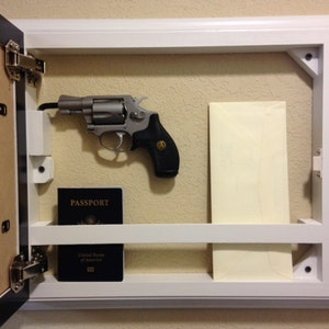 May include: A white framed wall-mounted gun safe with a hinged door. Inside, a silver revolver with a black grip is mounted. A dark blue passport and a cream-colored envelope are also visible.