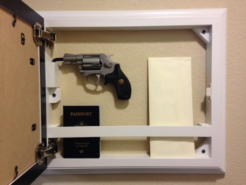 May include: A white framed wall-mounted gun safe with a hinged door. Inside, a silver revolver with a black grip is mounted. A dark blue passport and a cream-colored envelope are also visible.