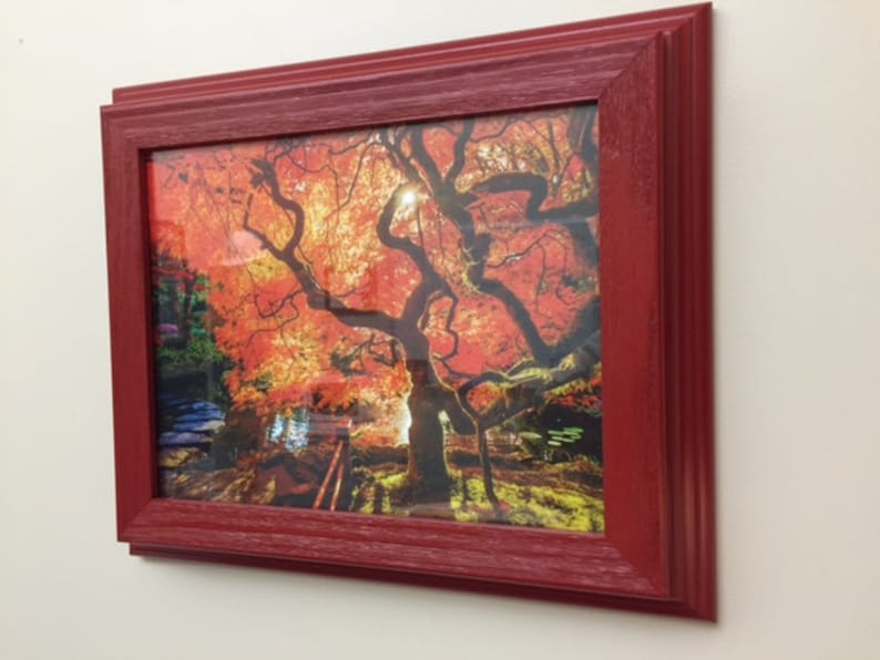 May include: A framed print featuring a vibrant autumn landscape with a large tree and red leaves. The frame is a deep red color, adding a bold contrast to the colorful scene. The artwork is displayed on a white wall.