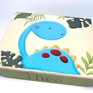 May include: A wooden box with a light green base and a light beige top. The top features a cartoon blue dinosaur with red spots and orange spikes, surrounded by green leaves. The name "Theo" is painted on the front of the box.