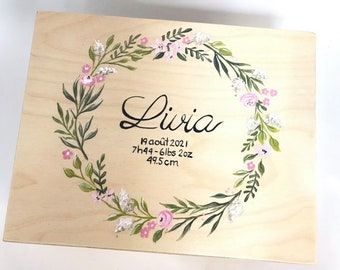 May include: A wooden box with a floral wreath painted around the edges. The wreath is in shades of green, pink, and white. The name "Livia" is written in cursive inside the wreath, along with the date "19 août 2021", the time "7h49", the weight "6lbs 2oz", and the length "49.5cm".