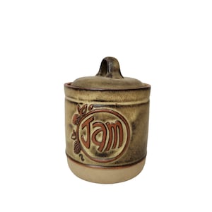 Może przedstawiać: Brązowy ceramiczny słoik z pokrywką i uchwytem. Słoik ma brązową i zieloną glazurę oraz brązowe koło z napisem "JAM". Wokół koła znajdują się małe brązowe jagody i liście.