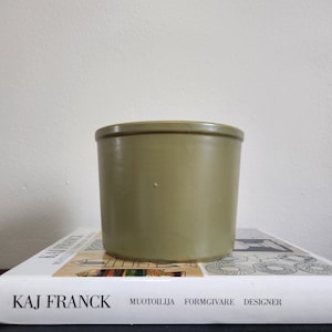May include: A green ceramic planter with a smooth, matte finish. The planter is sitting on a book with the text "KAJ FRANCK MUOTOIJA FORMGIVARE DESIGNER".