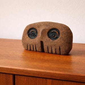 Puede incluir: Una taza de cerámica blanca con asa negra y el texto "But First, Coffee" en negrita negra. La taza se encuentra sobre un fondo borroso, sugiriendo un ambiente acogedor.