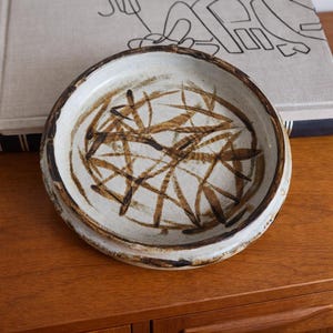 May include: A ceramic bowl with a wide rim and a cream-colored interior. The bowl features a brown, abstract design of intersecting lines. The rim is a darker brown, and the bowl sits on a wooden surface. The bowl is likely handmade.