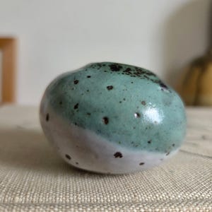 May include: A small, rounded ceramic vessel with a smooth, glossy finish. The upper portion is a soft mint green, while the lower section is a creamy white. The entire surface is speckled with small, dark brown dots throughout the glaze.