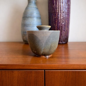 Può includere: Una ciotola in ceramica fatta a mano con coperchio, caratterizzata da un design asimmetrico unico. La ciotola ha una palette di colori verde e grigio tenui, con una superficie strutturata. Due vasi decorativi sullo sfondo aggiungono all'estetica. La ciotola è un'opera d'arte funzionale.