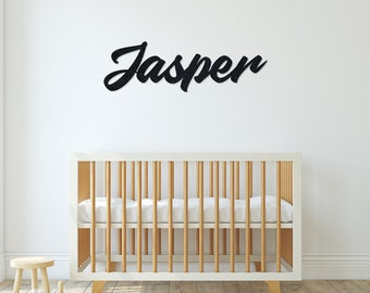 May include: A white crib with a wooden frame and a white mattress. The name "Jasper" is written in black cursive letters on the wall above the crib.