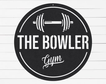 May include: Black metal sign with a white outline and text. The sign reads "THE BOWLER Gym" in a combination of block and script fonts. A barbell graphic is at the top of the sign, inside a circular shape. The sign is mounted on a white wooden background.