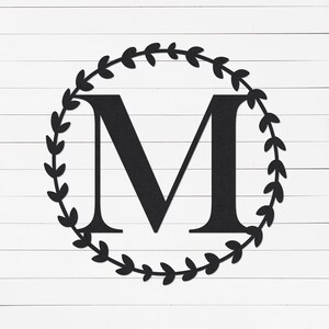 May include: Black metal letter "M" within a circular wreath of leaves. The letter is centred on a white wooden plank background. The wreath and letter are a solid black colour.